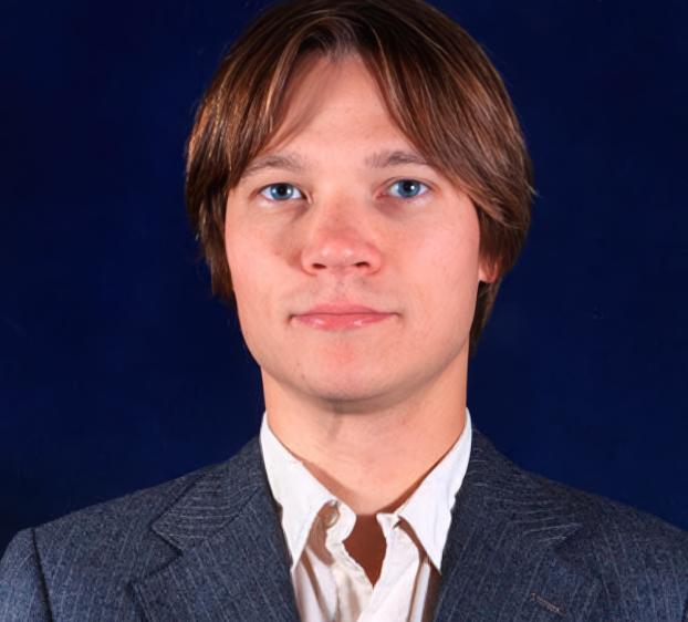 A head-and-shoulders portrait of a man with fair skin, bright blue eyes, and medium-length brown hair parted down the middle. He is wearing a dark, textured gray blazer over a white button-down shirt with an open collar. He has a neutral expression and is looking directly at the camera against a solid dark blue background.