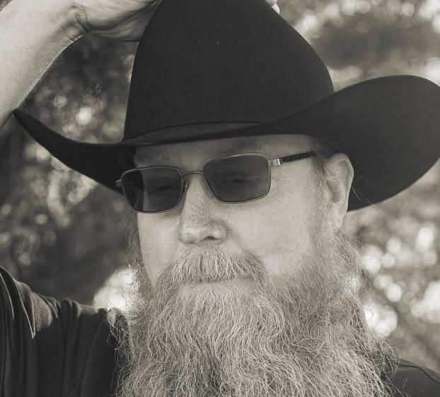 A black-and-white, close-up portrait of a man with a long, textured beard and mustache. He wears a dark cowboy hat, which he is adjusting with his right hand, and rectangular sunglasses. He is dressed in a dark polo shirt, and the background features softly blurred trees and natural light.