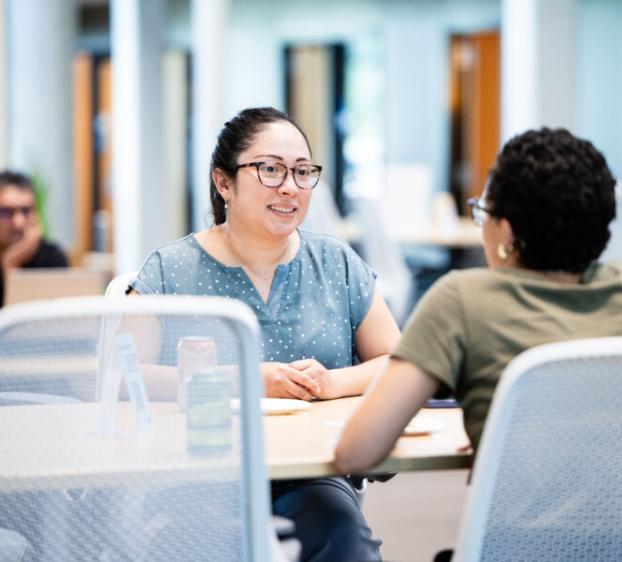 A woman with glasses and a blue polka-dot shirt sits at a table in a modern library, engaged in a friendly conversation with another person.