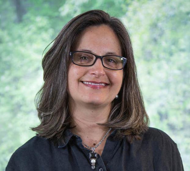 Headshot of a woman with shoulder-length brown hair and glasses, smiling at the camera. She is wearing a dark button-down shirt and a silver necklace with a black and white pendant. The background is a soft, out-of-focus green foliage.