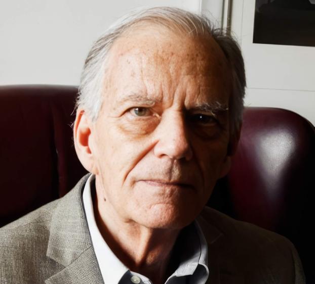 A close-up portrait of a man with light eyes and gray hair. He is wearing a grey textured blazer over a light blue button-down shirt. He sits in a dark reddish-brown leather chair against a neutral background. The lighting is soft, highlighting the features of his face as he looks directly at the camera with a neutral expression.