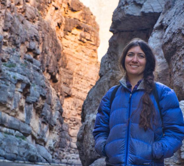 Woman wearing a blue jacket and backpack standing in a narrow rocky canyon, smiling at the camera with tall cliff walls rising on both sides.