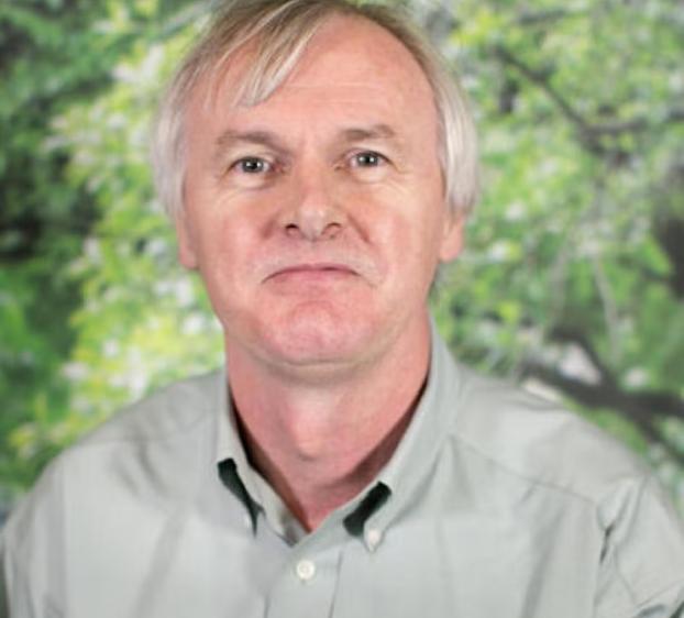 Headshot of a man with short light gray hair wearing a light green button-down shirt, looking at the camera with a neutral expression, set against a softly blurred green foliage background.