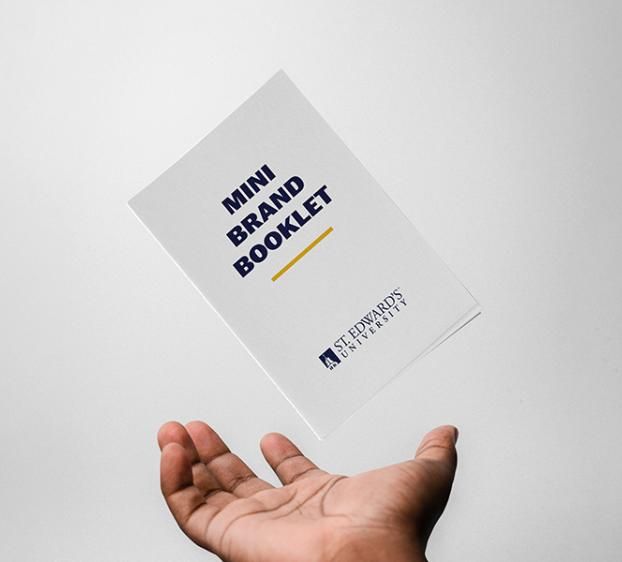 An open hand from below reaching up toward a small white booklet titled "MINI BRAND BOOKLET" with a yellow underline and the St. Edward's University logo. The booklet appears to be floating or tossed against a plain, light grey background.
