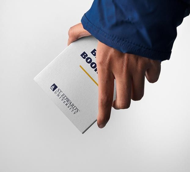 A person wearing a blue jacket holds a small white booklet featuring the St. Edward's University logo and a yellow accent line. The image is a close-up shot against a neutral, light grey background.