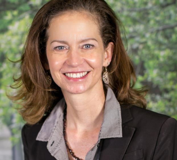 Smiling woman with shoulder-length brown hair wearing a dark blazer, gray shirt, and beaded necklace, photographed outdoors