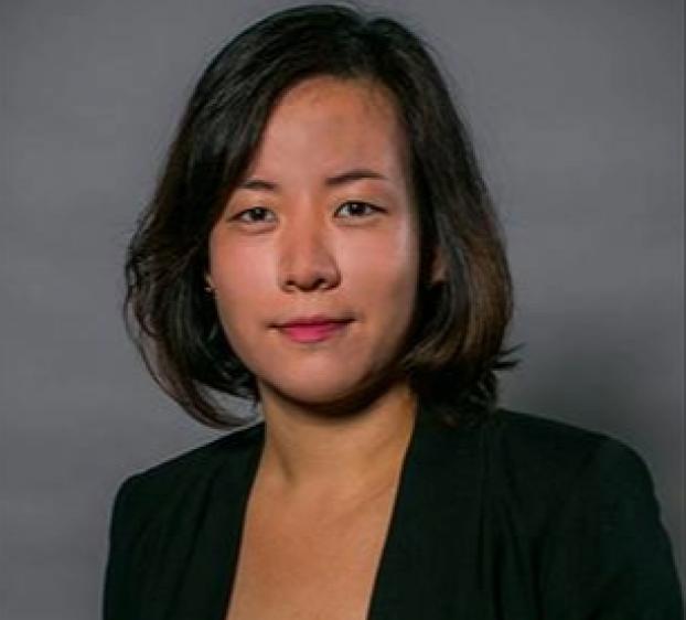 Portrait of a woman with shoulder-length dark hair wearing a black blazer over a dark floral top, posed against a gray studio background.