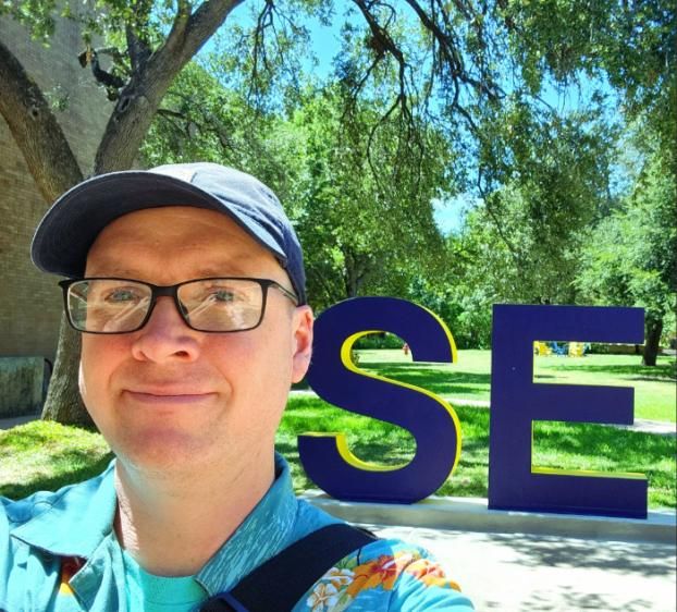 A headshot of a person wearing glasses, a blue cap, and a light blue shirt standing outdoors on a sunny day in front of large “SEU” letters, with trees and grass in the background.