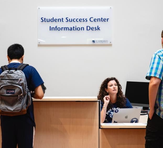 Two college students stand in front of a information desk and talk to a woman at a computer.