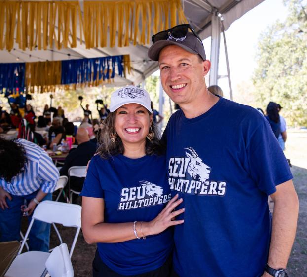 A woman and man wearing blue t-shirts smile for a photo outside.