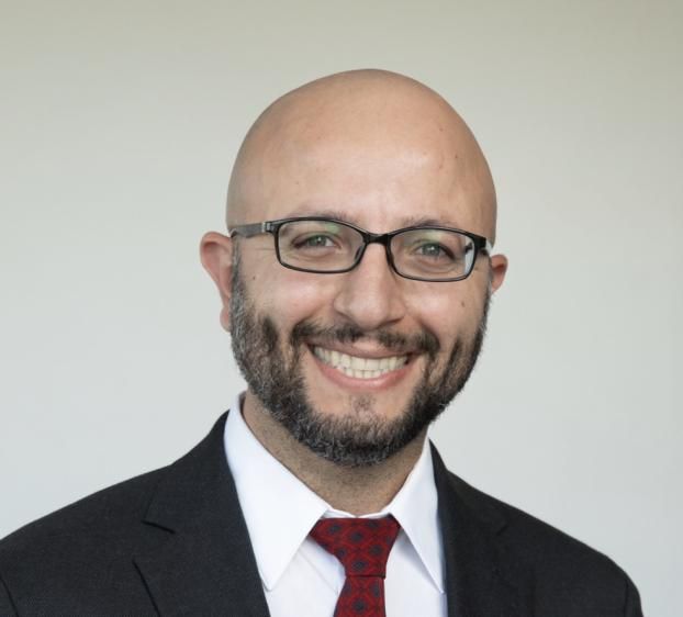 Professional headshot of a smiling bald man with glasses and a short beard, wearing a dark suit, white shirt, and red patterned tie against a light neutral background.