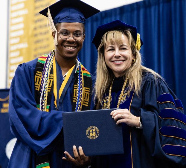 A graduate wearing a blue cap and gown smiles for a photo with a woman.