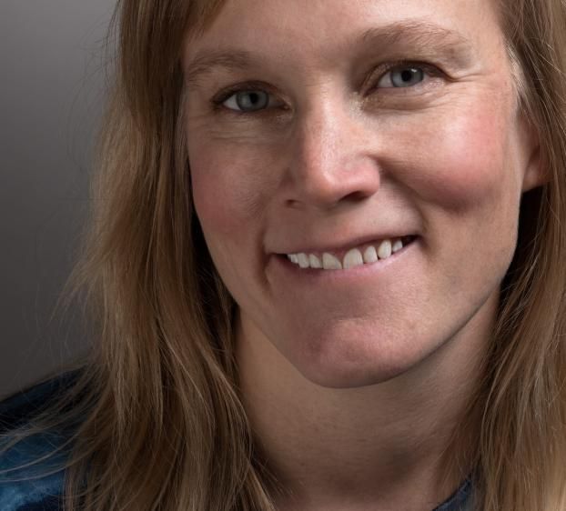 Close-up portrait of a fair-skinned woman with long blonde hair, smiling softly. She wears a blue patterned top, set against a neutral grey background with soft studio lighting.