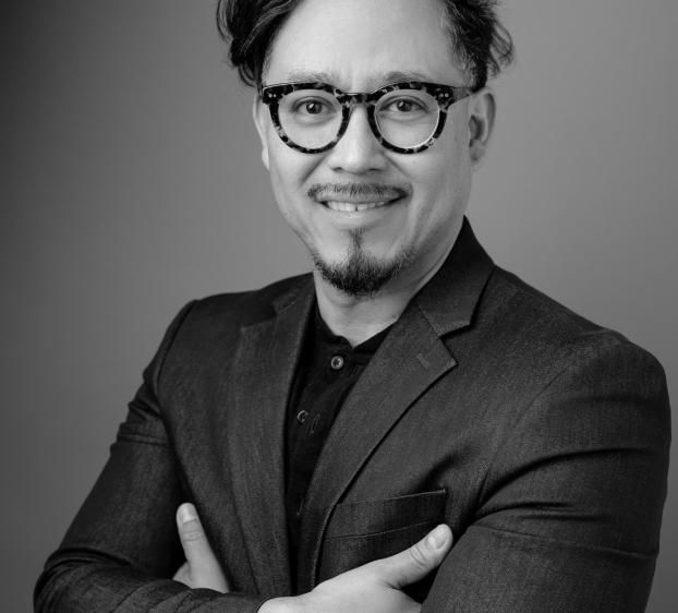Black-and-white studio headshot of a smiling man with arms crossed, wearing tortoiseshell glasses, a goatee, dark button-up shirt, and suit jacket against a plain backdrop.