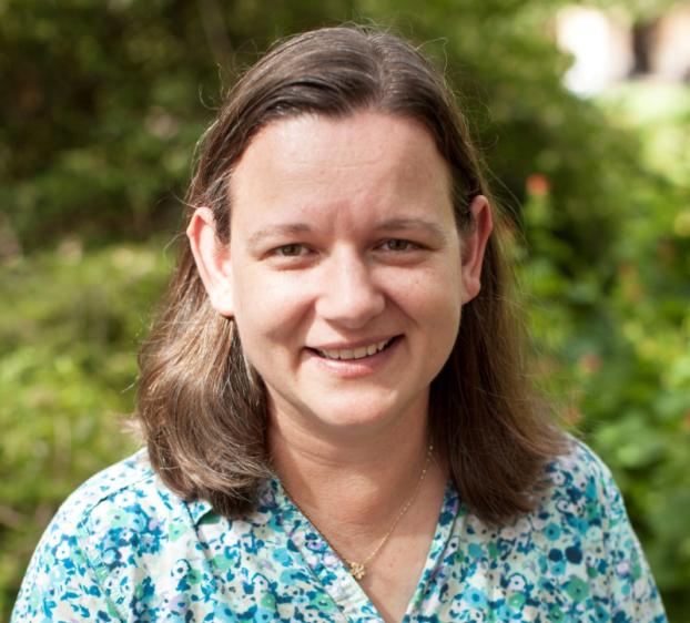 Head-and-shoulders portrait of a smiling woman with shoulder-length brown hair, wearing a blue and white floral blouse and a delicate necklace, photographed outdoors against a softly blurred green background.