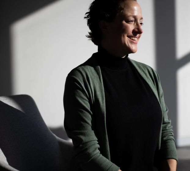 A woman with short dark hair smiling and sitting in a chair, illuminated by dramatic sunlight and casting strong shadows on a white wall behind her.