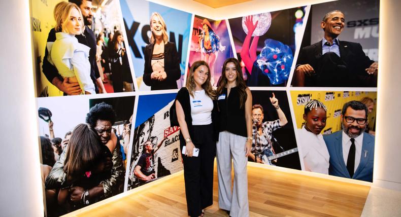 Students pose in front of a wall of photos from SXSW while visiting their offices in Austin for internships.