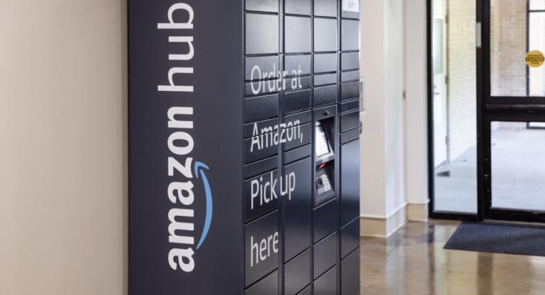 Amazon Hub Locker inside a building, featuring multiple secure compartments for package pickup. The dark-colored unit displays the text “Order at Amazon, Pick up here” and includes a small screen for user interaction. It is located in a hallway near glass doors leading outside.