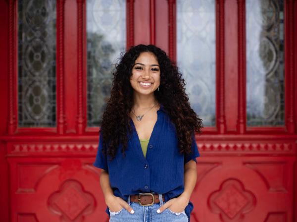 A young woman with curly hair stands smiling in front of an ornate red door with decorative glass panels. She is wearing a blue button-up shirt over a green top, paired with denim jeans. Her hands are in her pockets, and she is accessorized with a butterfly necklace. The vibrant red of the door contrasts with her outfit, creating a striking background for the portrait.