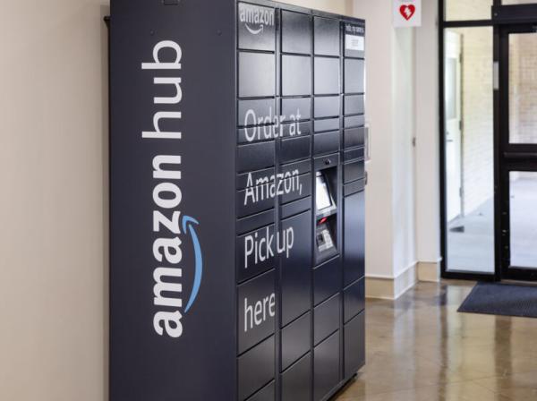 Amazon Hub Locker inside a building, featuring multiple secure compartments for package pickup. The dark-colored unit displays the text “Order at Amazon, Pick up here” and includes a small screen for user interaction. It is located in a hallway near glass doors leading outside.
