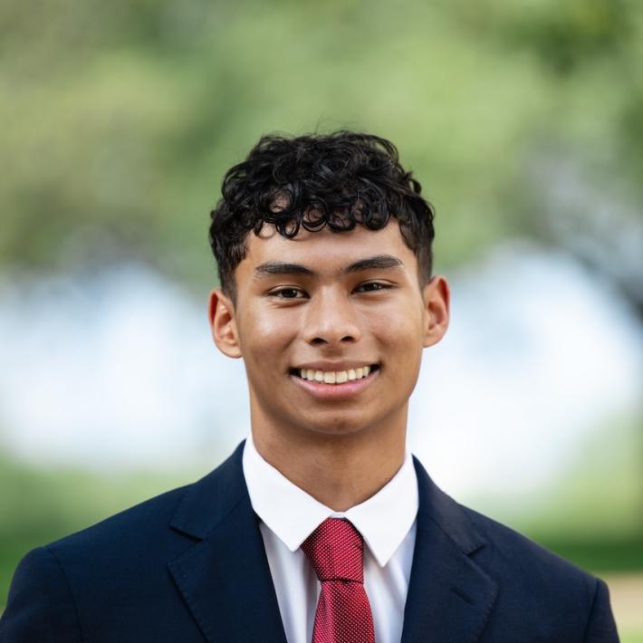 A young man wearing a navy suit, a white collared shirt and a red tie.