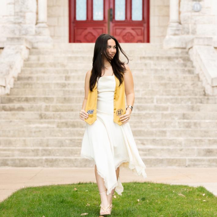 A young woman wearing a white dress and gold graduation stole walks across a lawn in front of an outdoor staircase and red doors.