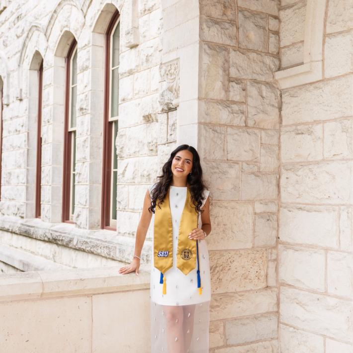A woman wearing a white dress and gold graduation stole smiles for a photo in front of a white brick building.