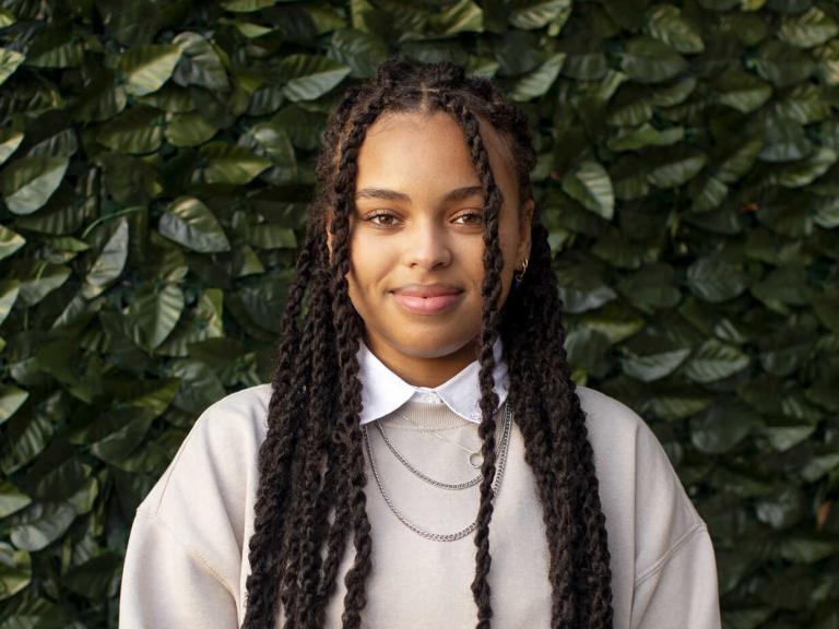 Taylor Huey wears a neutral tan sweatshirt with a white color shirt and multiple necklaces and stands in front of a green ivy background.