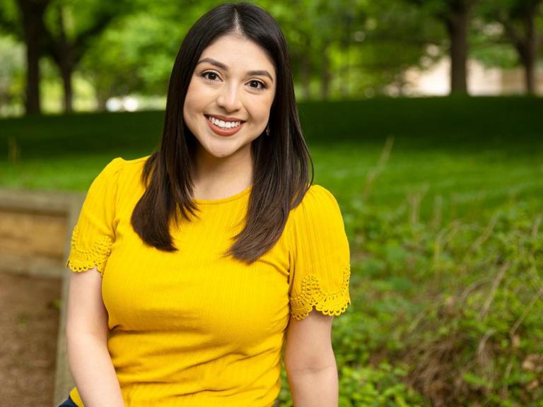 Victoria Garcia wears a yellow shirt and sits on a bench on Ragsdale patio. Greenery and trees are in the background.