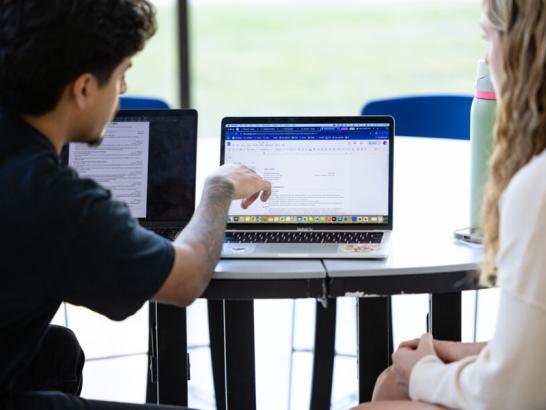A man and a woman sit at a table together and look at a computer screen, which displays a document that the man is pointing to
