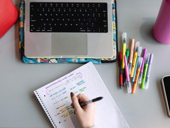 A student takes color-coded notes on a paper notebook. A laptop, water bottle, pens, cell phone and bag are also seen in this overhead scene.