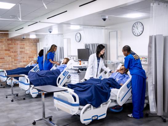 Nursing students and a faculty member work in a nursing simulation lab. The students wear blue scrubs and the faculty member wears a white coat. They interact with mannequins in mock treatment.
