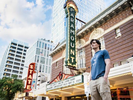 A student wears a grey-blue shirt and khaki pants and stands in front of the Paramount and State Theatres. The photo is taken at a low angle so as to see more of the downtown Austin skyline.