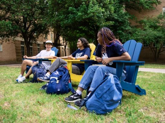 Three students sit in blue and yellow wooden chairs outside a residence hall, with trees in the background. Their backpacks are by their chairs as they sit and chat with one another.