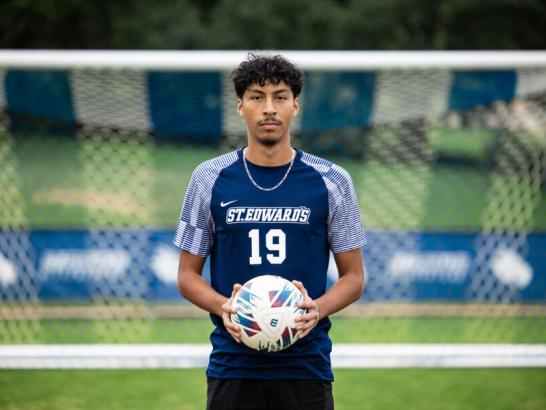 A soccer player stands on the field and holds a soccer ball. The goal is blurred in the background. He wears his blue and white athletics uniform.