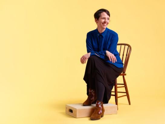 A woman wearing a blue blouse, black skirt and brown boots sits on a wooden chair with one of her feet on a wooden block. She has her arms crossed in her lap and she smiles as she looks away from the camera. She sits in front of a warm yellow backdrop.