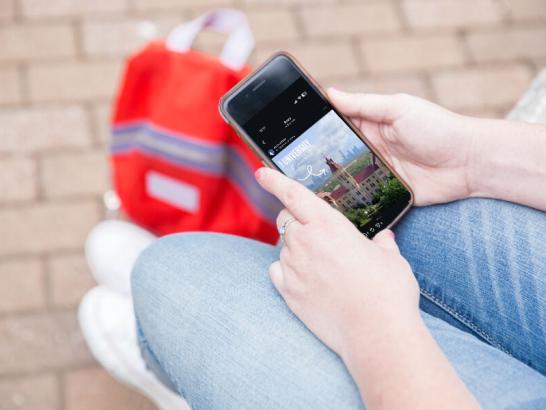 A person is shown from an aerial view sitting down, with only legs and hand showing, holding a phone and resting a red backpack at their feet