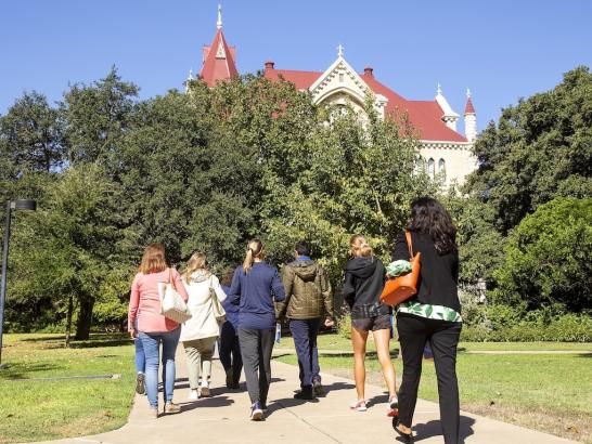 A group of prospective students and families walk along a sunny concrete path toward the historic, red-roofed Main Building at St. Edward's University. Lush green trees line the walkway, partially framing the gothic limestone architecture under a clear blue sky. The shot is from behind the group, capturing a vibrant day of campus exploration.