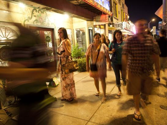 A busy nighttime street scene on South Congress Avenue in Austin, featuring pedestrians walking along a sidewalk lined with eclectic shops like Uncommon Objects. The image uses a long exposure, creating motion blur for several people in the foreground and background. Warm, glowing storefront lights illuminate the sidewalk, capturing the vibrant and energetic atmosphere of the local district.