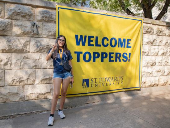A young woman in a navy t-shirt and denim shorts stands next to a large yellow banner that reads "WELCOME TOPPERS! ST. EDWARD'S UNIVERSITY." She is smiling and making a Toppers Up hand gesture against a light-colored stone wall.