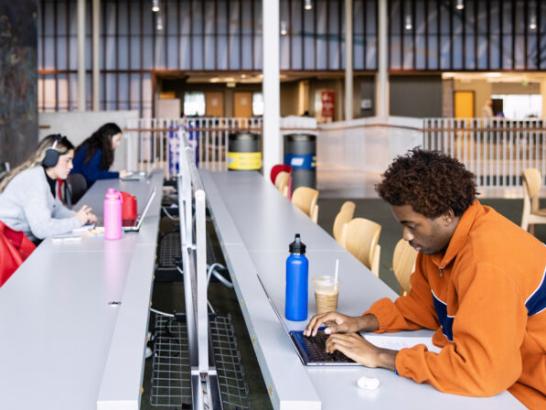 Students study at long gray tables in a modern, open-concept library with high ceilings. In the foreground, a young man in an orange pullover types on a laptop; across from him, several other students work on computers with various water bottles and headphones.