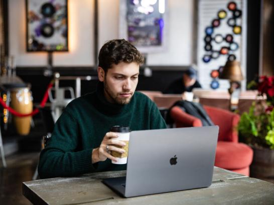 A young man in a dark green turtleneck sits at a cafe table, focused on his laptop while holding a coffee cup. The background is softly blurred, showing a cozy interior with musical decor and a red armchair.