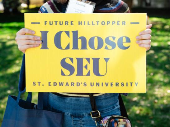 A smiling young woman stands outdoors, holding a bright yellow sign that reads "Future Hilltopper - I Chose SEU - St. Edward’s University." She is wearing a name tag and has a blue tote bag over her shoulder, with a backdrop of sunlit trees.