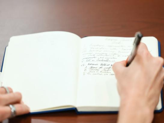A person’s hands are shown writing in a small, blue notebook with a silver pen on a dark wooden surface. The notebook contains handwritten notes and the person is wearing rings on their left hand, which holds the notebook open.