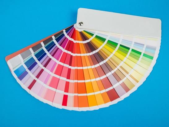A Pantone color swatch book fanned out to display a wide spectrum of colors, including reds, oranges, yellows, greens, and purples, set against a solid bright blue background.