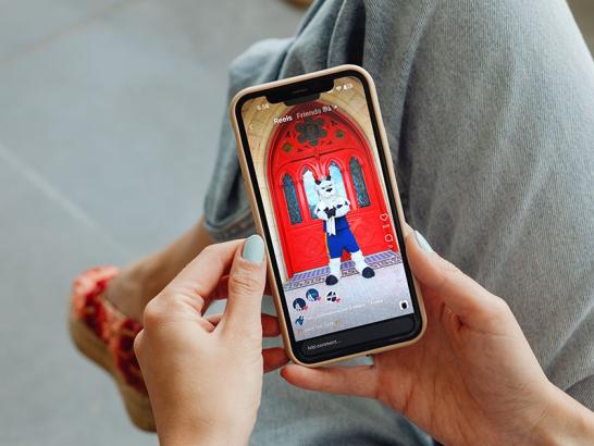 A top-down view of a person holding an iPhone, scrolling through Instagram Reels. The phone screen displays a video of the Topper goat mascot standing in front of large, ornate red doors. The person has light blue nail polish and is wearing denim jeans and patterned shoes.