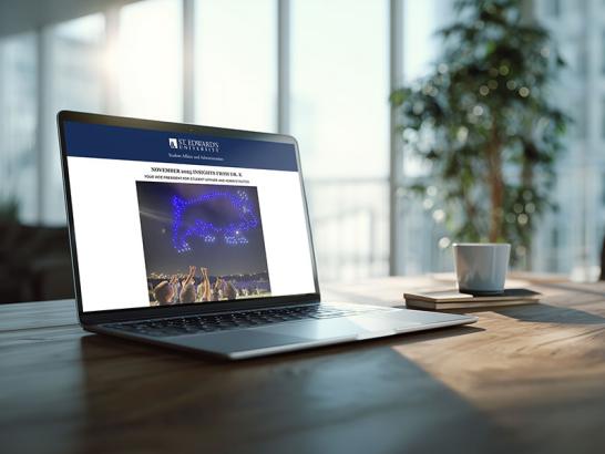 A laptop sits on a wooden desk in a bright, modern office, displaying an email newsletter from St. Edward's University titled "November 2025 Insights from Dr. K." The screen shows a photo of a drone light show forming the shape of a goat in the night sky.
