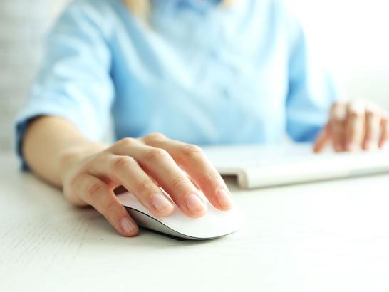 A close-up shot of a person's hand resting on a white, modern computer mouse. In the soft-focus background, the person is wearing a light blue shirt and has their other hand near a keyboard on a white desk.