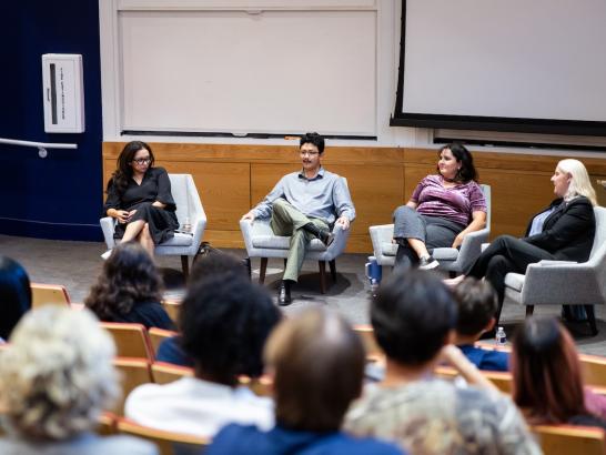 Three women and one man sit on a panel in front of a stage. 