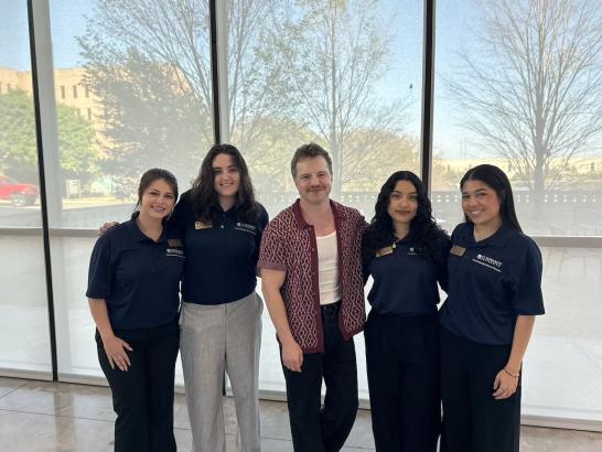 A group of female college students smile for a photo with a guest speaker in front of a window.